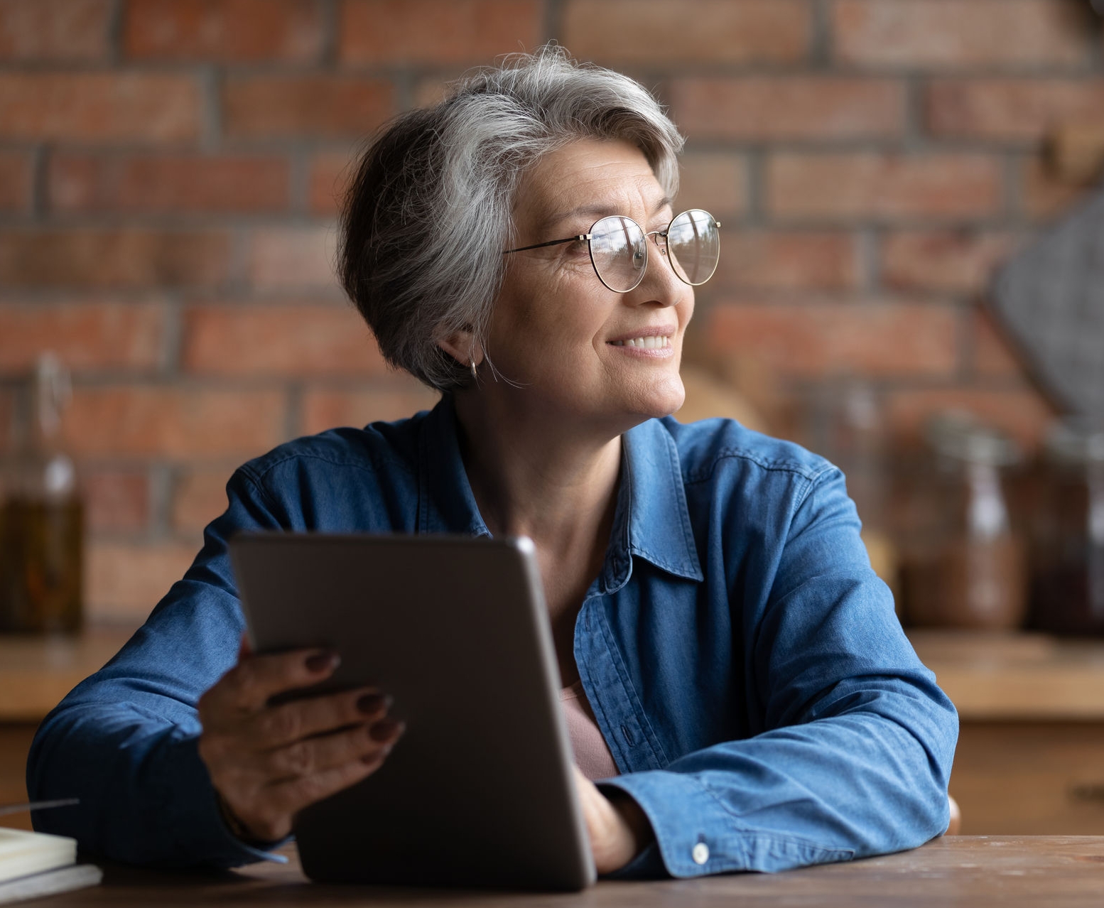 Dreamy senior grey haired woman in spectacles holding digital computer tablet in hands, looking in distance, copy space for advertising text of retired old people and modern technology easy usage.