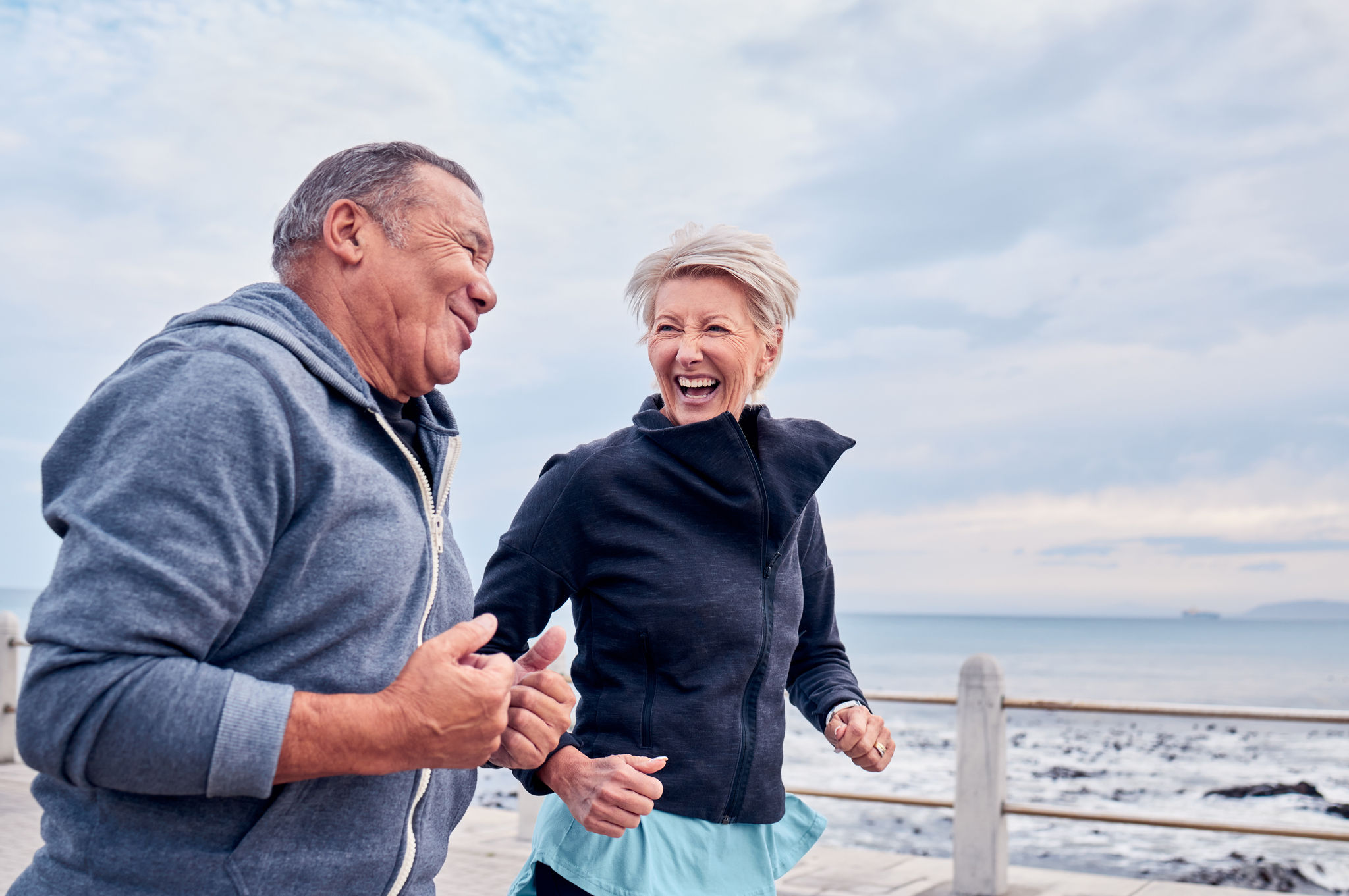 Running, fitness and senior couple laugh by ocean for exercise, healthy body and wellness in retirement. Sports, marriage and happy elderly man and woman for smile, cardio workout and training by sea.